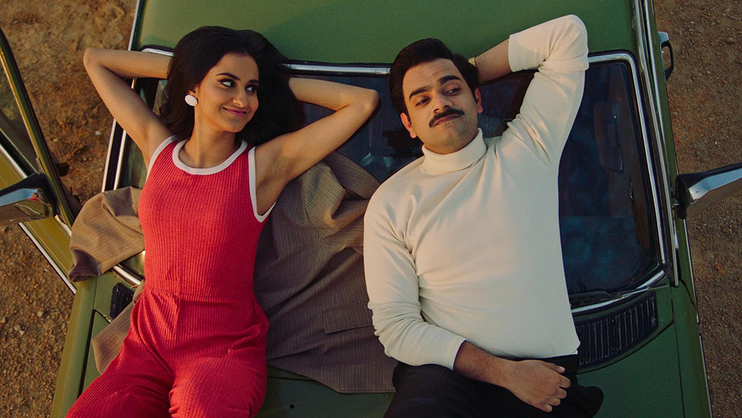 A vibrant scene from 'The Queen of My Dreams' showing Mariam and Azra, draped in colorful Bollywood-inspired attire, lost in a moment of laughter and music.